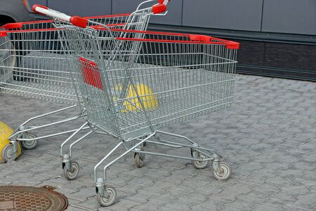 two empty gray metal empty trolleys with red handles on wheels stand on the sidewalk on the street...