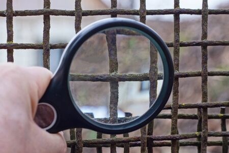 black round magnifier in the hand magnifies the brown rusty iron grate in the old wall