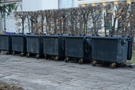 a row of black plastic garbage cans stand on gray asphalt near dry trees in a city park