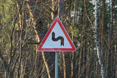one triangular road sign dangerous turn on a pillar against the background of trees on the streetの写真素材