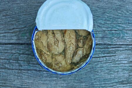 one open white tin can with brown pieces of fish in oil stands on a gray wooden tableの写真素材