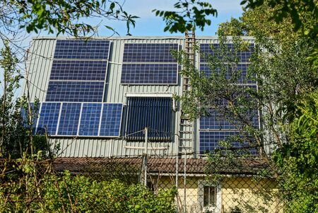gray slate roof of a private house with blue solar panels against the sky and green vegetation