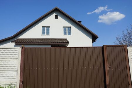 brown metal gate and part of a concrete fence in front of the facade of a white private house against a blue skyの写真素材