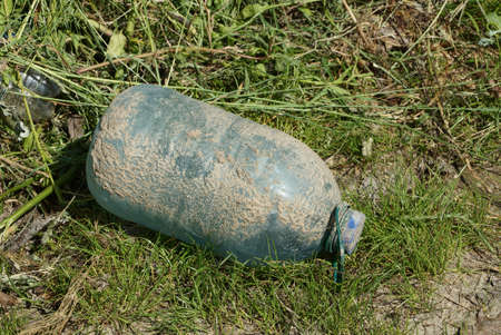 trash from one big blue plastic bottle in gray mud on green grass in nature