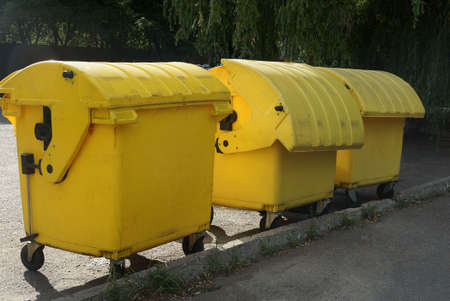 three yellow closed trash cans stand on the gray asphalt outside