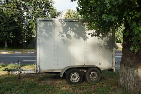 gray metal trailer trailer on black wheels stands on the street in the green grass by the tree