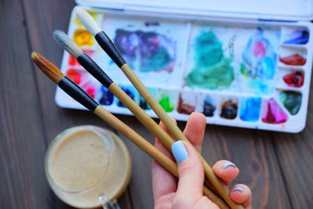 three brushes in the artist's hand on the background of a box with colored paints and a glass cup...