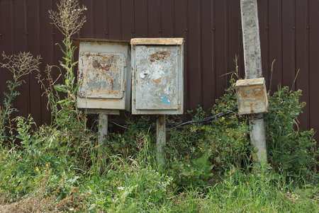 three gray old metal boxes for electrical wires in green grass outside near a brown wallの写真素材