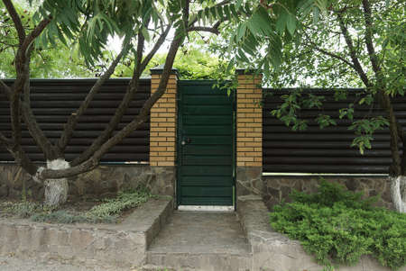 closed iron green door and brown wall of a fence made of bricks and wooden planks outside with decorative treesの写真素材