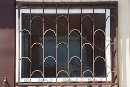 one large square window behind a brown iron grating on the wall of a building on the street