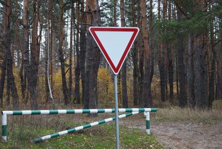one triangular road sign give way near a striped closed barrier against a background of pine trees