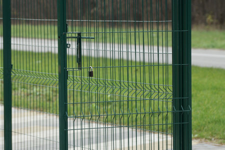 part of a green fence made of metal rods in a net and a open door on a gray sidewalk and grass near...