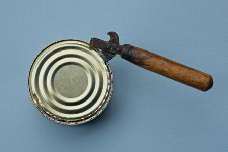 an old can opener with a brown wooden handle opens a yellow aluminum lid on a tin can on a gray...