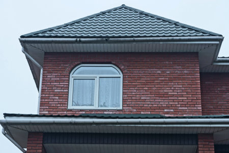 the attic of a brown brick house with a large window under a green tiled roof against the sky