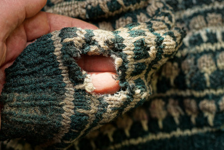 fingers are holding black brown woolen fabric of an old sweater with a large torn hole