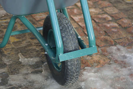 a black wheel on a green metal garden wheelbarrow stands on a brown sidewalk and white snow on a...