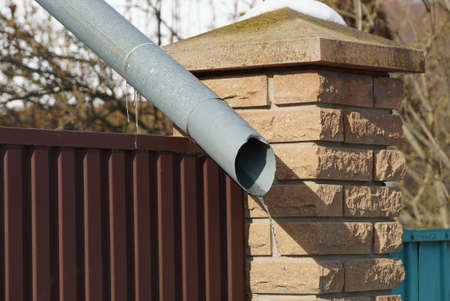 long gray metal gutter pipe on a brown wall of a fence made of iron and bricks outside