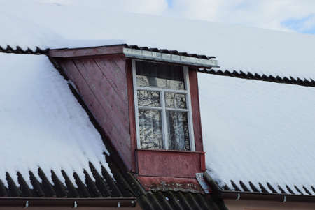 red wooden attic with a window on a gray tiled roof in white snow against the skyの写真素材
