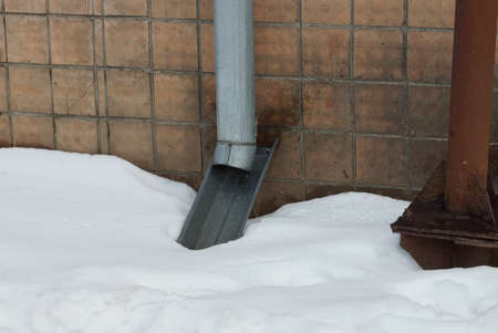 gray metal gutter pipe on a brown wall of a house above a snowdrift of white snow outsideの写真素材