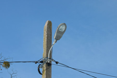 gray iron lantern with a glass bulb on a concrete pole with black electric wires against a blue skyの写真素材