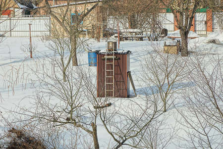 rustic brown building a summer shower with a ladder and a metal tank stands in the winter garden among gray trees and white snowの写真素材