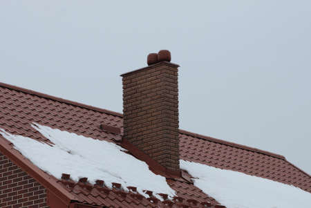 one big brown brick chimney on a red tiled roof of a private house in white snow on a winter street against a gray skyの写真素材