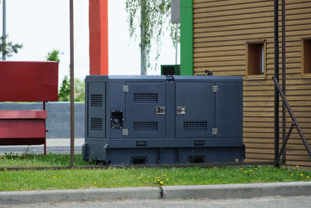 one black iron generator stands in green grass on the street against a brown wall