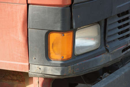 one front large glass headlight on the cab of a red black truck on the streetの写真素材