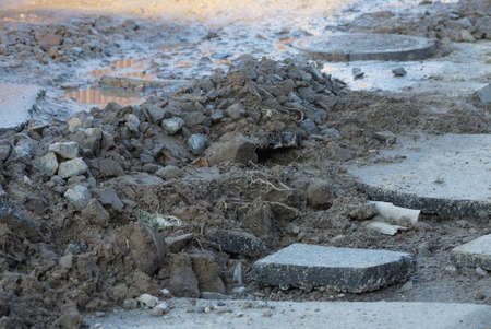 part of a repaired road with a pile of gray earth and stones near pieces of asphalt in wet mud