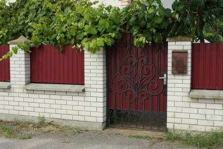 closed red metal door with a black wrought iron pattern and part of a white brick wall of a fence...