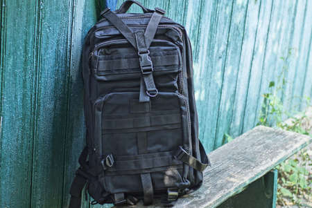 one black tactical backpack stands on a gray wooden bench against a green wall on the streetの写真素材