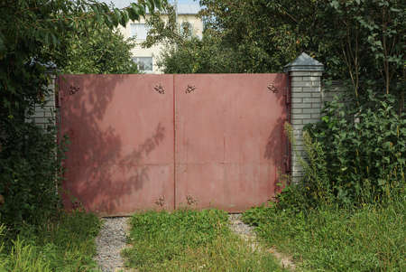 one closed red metal gate and a white wall of a brick fence overgrown with green vegetation and grass on the streetの写真素材
