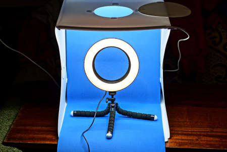 a white plastic lightbox with a blue background and a selfie ring lamp on a tripod stand on a brown table on a black backgroundの写真素材