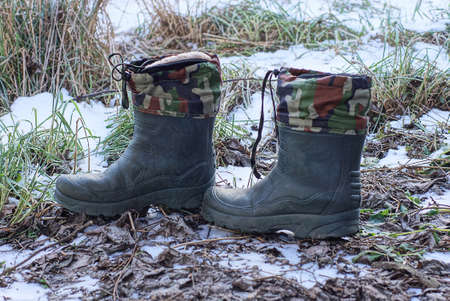two winter boots made of green rubber and spotted fabric stand on brown ground in grass and white snow on a winter streetの写真素材