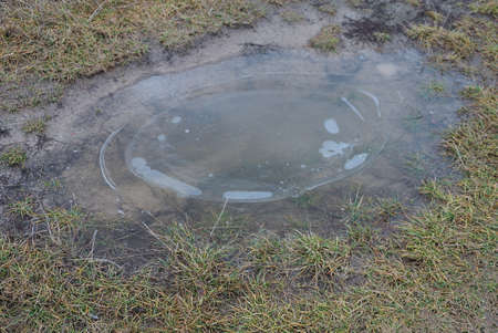 part of a rural road of gray earth with a lone pothole and frozen water in the ice in green grass