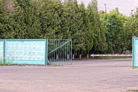 an open green iron gate and a white barrier on the street on gray asphalt near the concrete wall of...