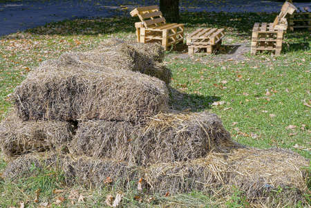 a pile of bales of gray dry hay on green grass in a park near a wooden table and benchesの写真素材