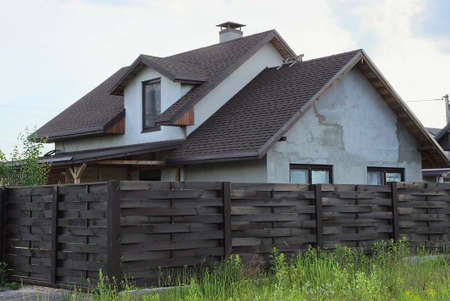 one gray private house under a brown tiled roof behind a wooden fence in green grass against the skyの写真素材