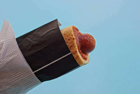hot dog food with sausage with red ketchup in bread in black paper packaging and a white napkin on a blue backgroundの写真素材
