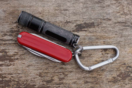 a black small flashlight and a red folding knife on a gray iron carabiner latch lies on a gray...