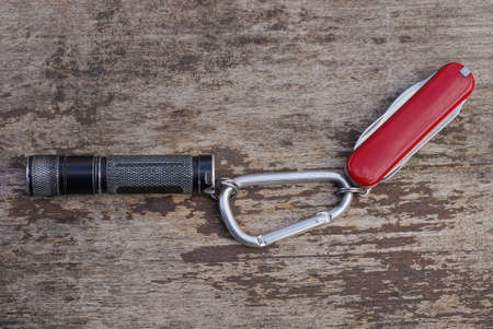 a black small flashlight and a red folding knife on a gray iron carabiner latch lies on a gray...