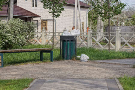 one large bin full of rubbish stands in the green grass near the gray sidewalk and an empty park bench near the white wooden fenceの写真素材