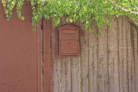 one brown metal mailbox hanging on a gray wooden fence wall in the street in green leaves on the...