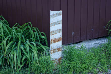 one iron striped boundary post stands on the street against a brown metal wall of the fence in green...