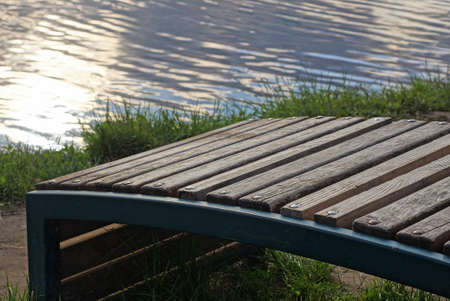 one empty brown wooden bench stands in green grass by a earth in a park on the shore of the lake with gray waterの写真素材