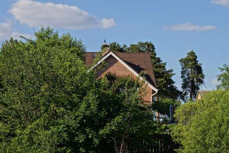 brown brick attic of a private house under a tiled roof in green trees against a blue sky