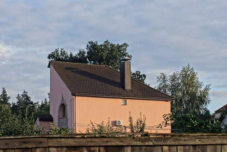 one brown private house under a tiled and a metal chimney on the street among green trees against the sky and gray clouds behind a wooden roof fenceの写真素材