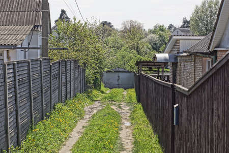 rural alley with a road in green grass along fences made of gray concrete and brown wooden boards and gray metal gates in a dead endの写真素材