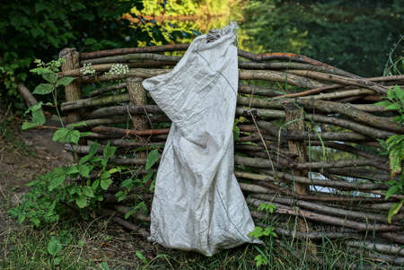 one white garbage bag hanging on a brown wooden fence made of branches on a street in green vegetationの写真素材