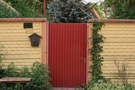 one red closed metal door on a brown concrete fence wall on a rural street in green vegetationの写真素材